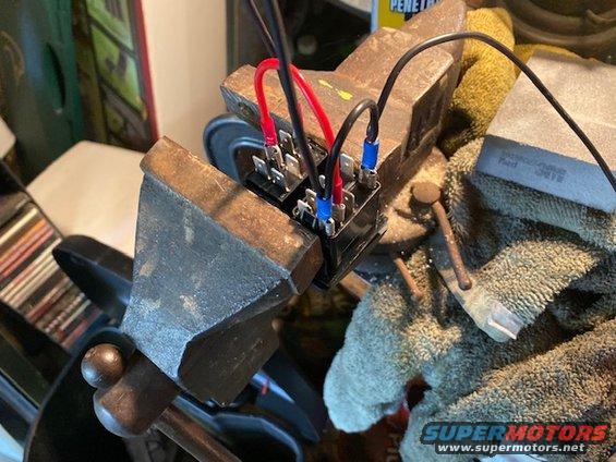 Two automotive relays with red and black wires attached, held in a metal vise