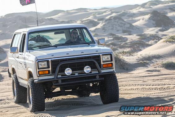 White 1981 Ford Bronco driving on sand dunes with a pink flag