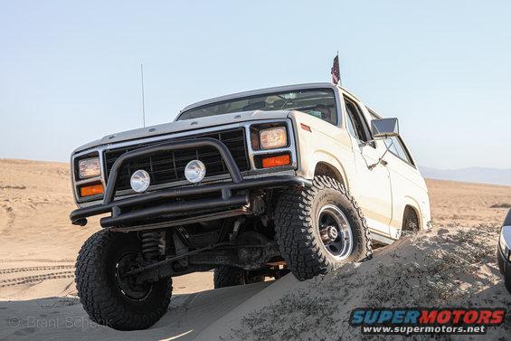 White Ford Bronco with bull bar and off-road tires driving on sand dunes. Another vehicle is partially visible on the right