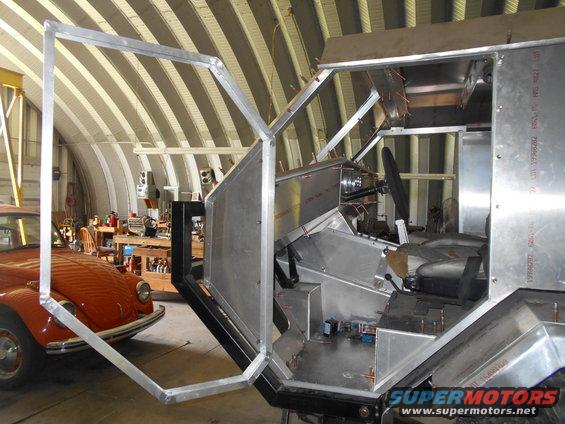 Custom vehicle interior with exposed metal panels, seats, and steering wheel. An orange car is visible to the left
