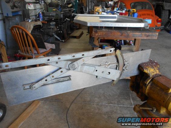 Window regulator mechanism clamped in a vise, with a wooden chair and orange car in the background