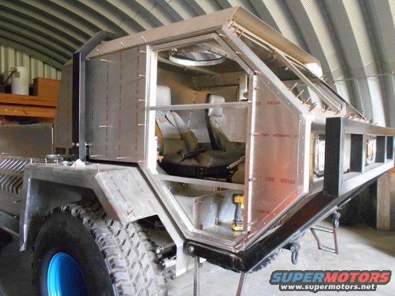 Unfinished custom vehicle with metal body panels and exposed interior, showing seats and a drill. It has large off-road tires