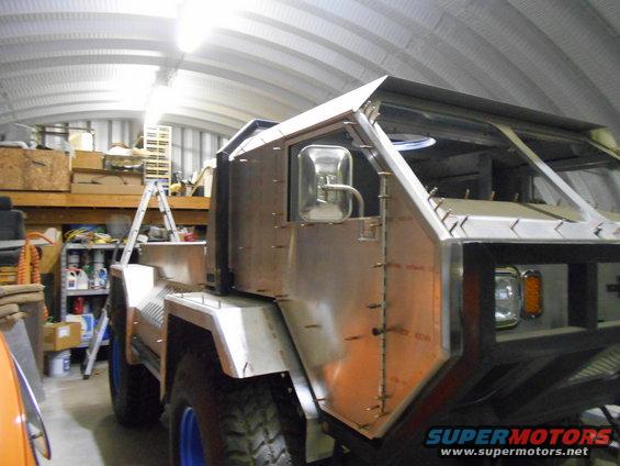 Unfinished custom truck with aluminum body panels and blue wheels, viewed from the front-right