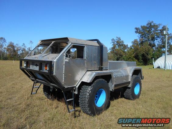 Custom-built off-road truck with a silver metal body and bright blue wheel rims, parked in a grassy field