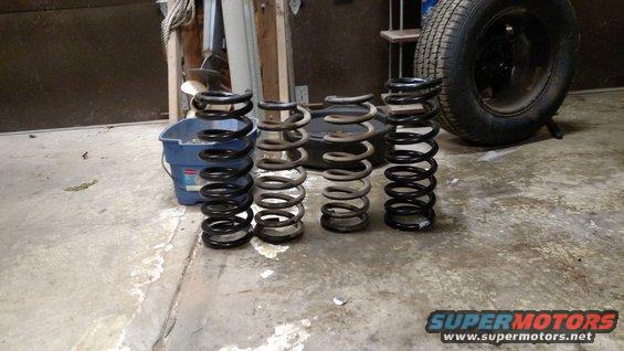 Four coil springs of varying sizes and conditions sit on a concrete floor next to a blue bucket and a tire