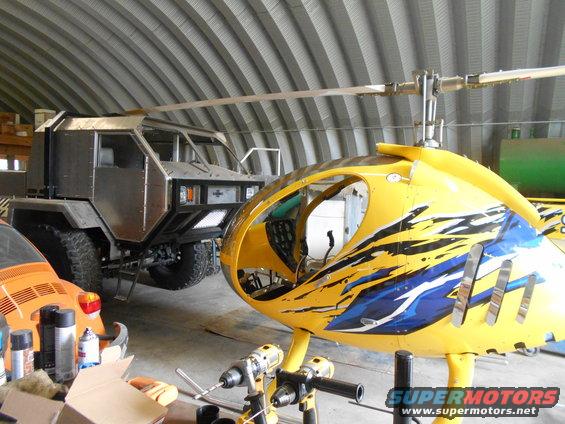 Custom armored vehicle and yellow helicopter fuselage in a workshop. Orange VW Beetle partially visible on left
