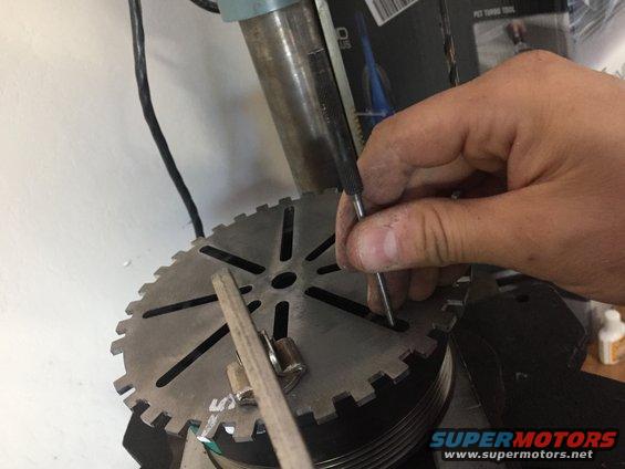 Hand using a scribe on a large toothed metal disc, possibly part of a drill press