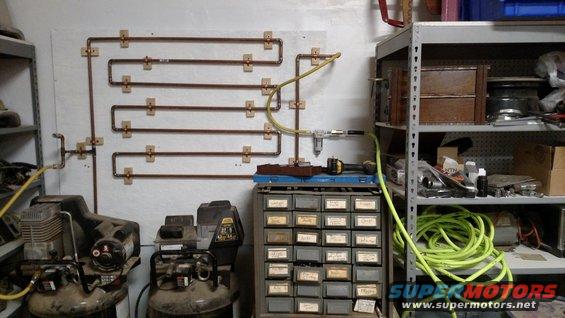 Two air compressors sit below a wall-mounted copper pipe system. A metal cabinet with small drawers is nearby, along with shelves holding various items