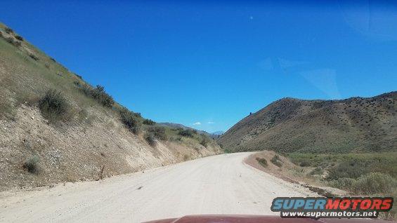 Dirt road winding through dry, grassy hills under a clear blue sky. Part of a red vehicle hood is visible at the bottom