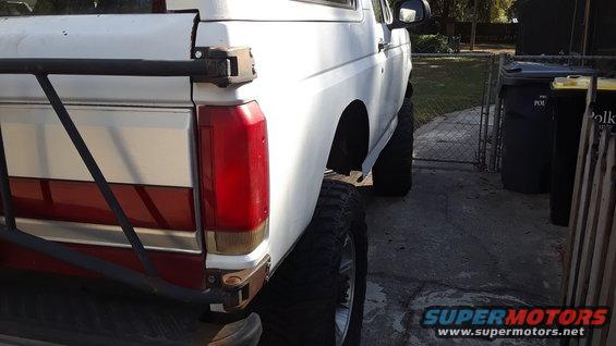White Ford Bronco with red stripe, lifted suspension, and aftermarket rear bumper. Visible from the rear quarter panel
