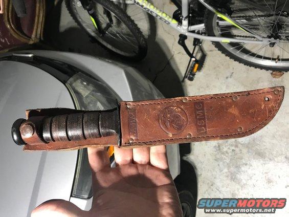 Hand holding a USMC combat knife with a leather sheath stamped with USMC emblem and text