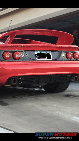 Red sports car rear with V8 badge, quad exhaust pipes, and large spoiler. License plate obscured