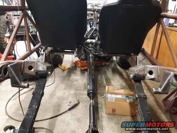Two black bucket seats in a disassembled vehicle chassis with leaf springs and a driveshaft visible