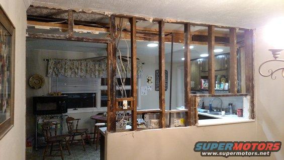 Interior view of a kitchen undergoing demolition, exposing wall studs and wiring. A microwave and chairs are visible in the adjacent room