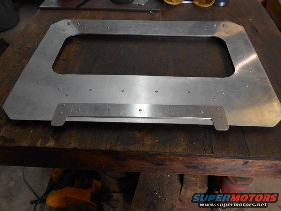 Aluminum frame with a rectangular opening, mounted on a wooden workbench. It has numerous small holes drilled around the opening and along a lower horizontal bar