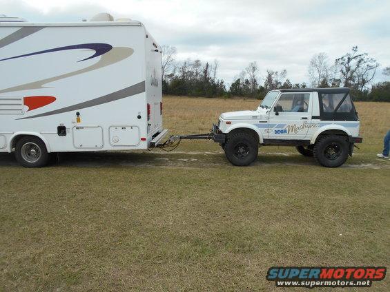 White off-road vehicle with black soft top towing a white RV, side view