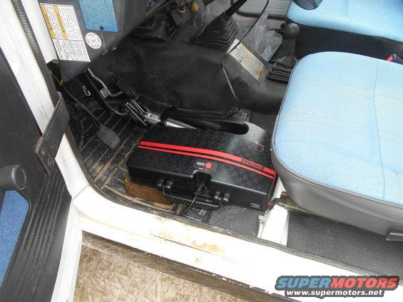 Interior view of a vehicle showing the driver's side floor, pedals, and a black portable brake system with red stripes. Blue seats are visible