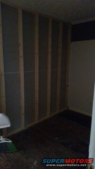 Interior framing of a room with wooden studs and dark wood floor. Broom and dustpan visible