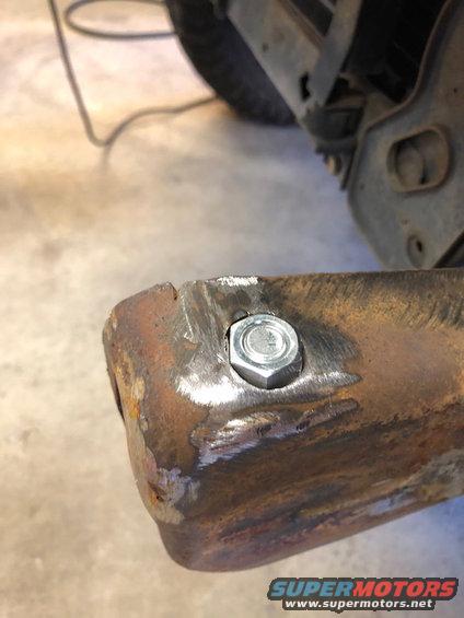 Rusty metal bracket with a new bolt and nut, showing weld marks and a tire in the background