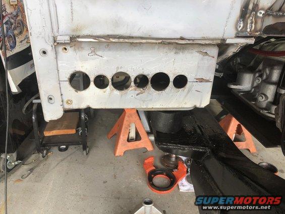 Vehicle frame section with multiple circular cutouts, supported by jack stands. Some engine components visible