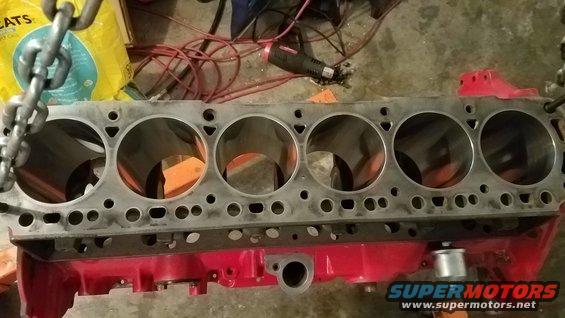 Engine block with six cylinders, suspended by a chain. Tools and a bag are in the background