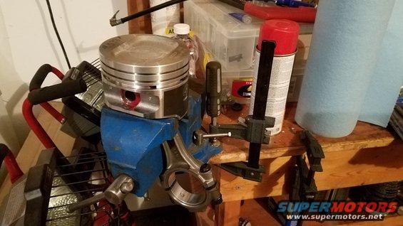 Piston and connecting rod clamped in a blue vise on a workbench with tools and supplies in the background