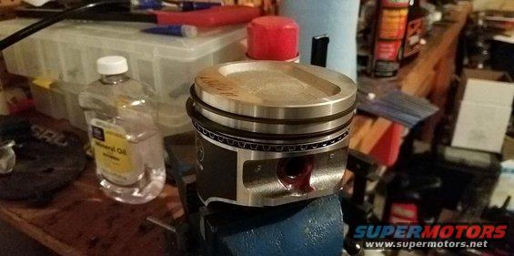 Piston held in a blue vise on a workbench, with a bottle of mineral oil and various tools in the background