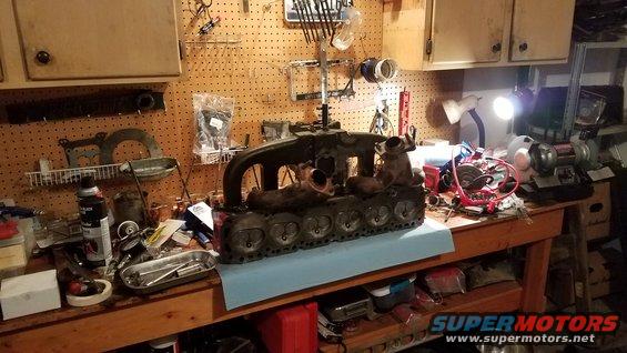 Engine block with exhaust manifolds attached, resting on a workbench with tools and parts