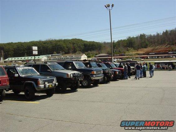 Line of SUVs and trucks parked in a lot with people standing near the rear