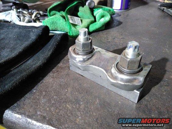 Metal bracket with two large bolts and nuts, resting on a workbench next to black fabric and green cloth
