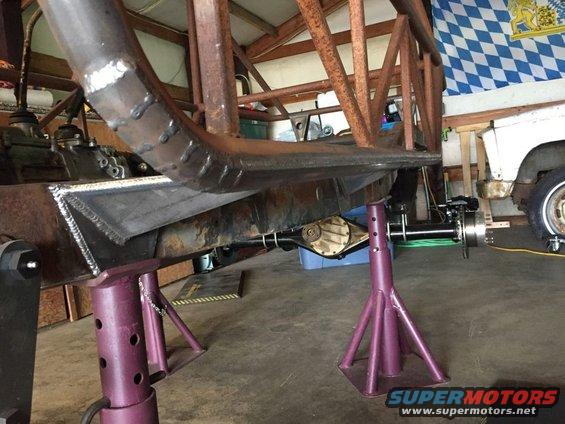 Vehicle frame on jack stands with suspension components visible. Another vehicle is partially seen in the background