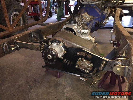 Vehicle chassis with transmission and transfer case, supported by jack stands. A large tire is visible on the left