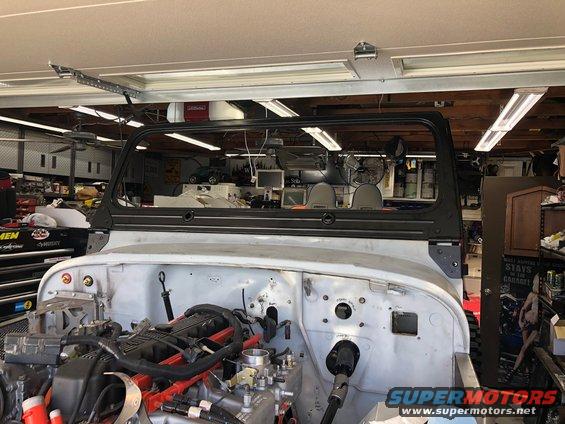Front end of a Jeep Wrangler body, engine exposed, in a garage with tools and shelves in background