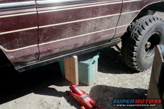 Maroon vehicle side with white pinstripes, lifted by a red jack and green bin, showing a large off-road tire