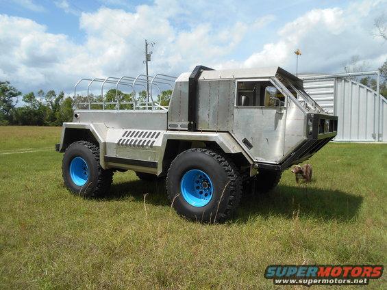 Silver custom off-road vehicle with blue wheels, metal body, and roll cage in a grassy field
