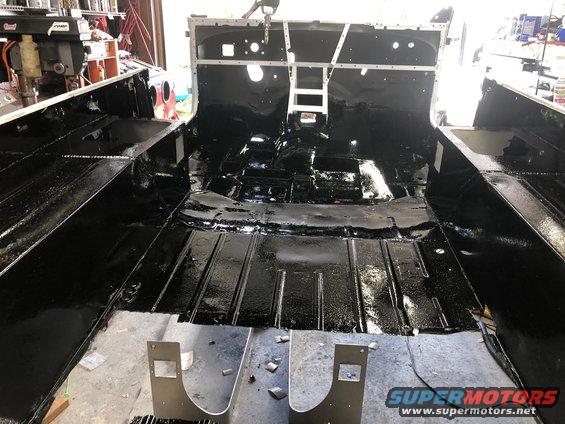 Interior of a Jeep CJ with black undercoating applied to the floor pans and body tub. Some metal brackets are visible in the foreground
