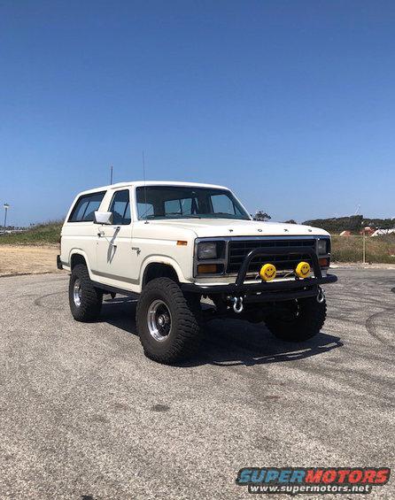 White Ford Bronco with large tires and a brush guard, parked on asphalt under a clear blue sky