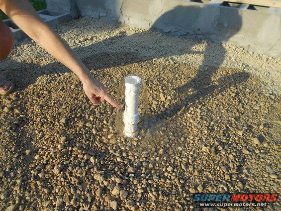 hand pointing at a white pipe sticking out of gravel, with a concrete structure in the background