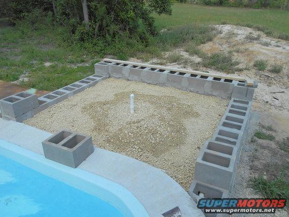 Construction site with cinder blocks forming a rectangular base, gravel fill, and a white pipe in the center, next to a blue pool