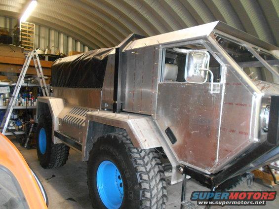 Custom truck build with aluminum body panels, large off-road tires with blue rims, and a black canvas top. Ladder and shelves visible in background
