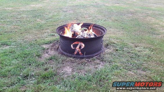 Fire pit made from a tire rim with a cutout 'R' logo, burning wood in a grassy yard