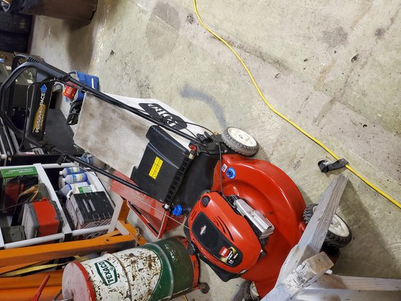 Two lawnmowers and old gas cans sit on a concrete floor with a yellow cord snaking nearby
