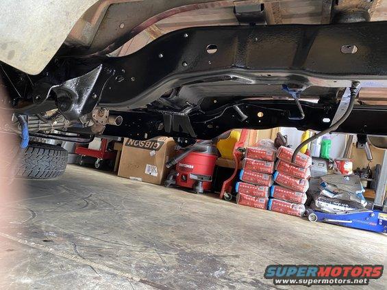 Underside of a vehicle frame with black paint, showing suspension components and a tire. Boxes and tools are visible in the background