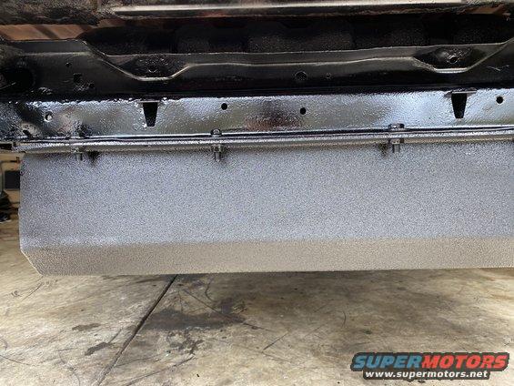 Close-up of a vehicle's underbody showing a gray textured panel and black frame components. Supermotors.net logo visible