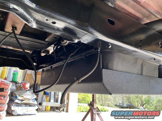 Underside view of a vehicle frame with a newly painted fuel tank and brake lines. Some bags of concrete and cleaning supplies are visible in the background