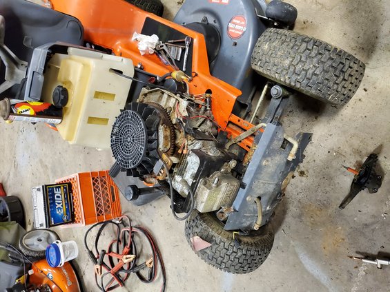 Orange lawn mower engine and parts on floor. EXIDE battery visible