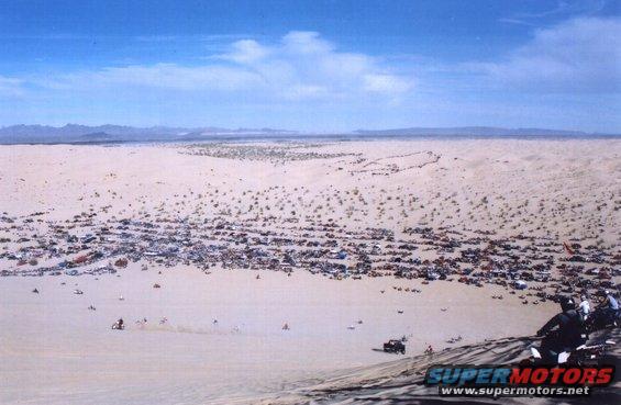 Desert scene with numerous parked vehicles and people riding ATVs on sand dunes under a blue sky