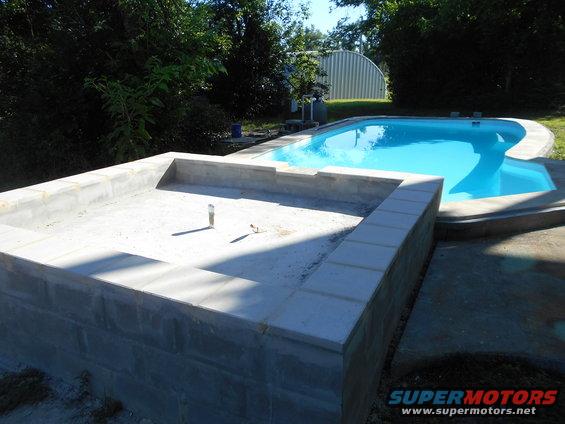 Under construction swimming pool with a separate shallow area, surrounded by trees and a metal building in the background
