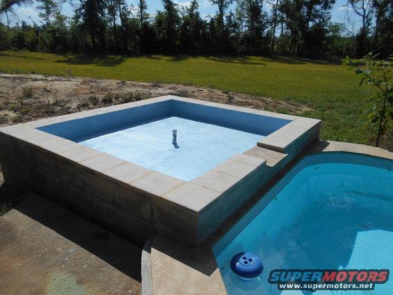 Two empty swimming pools with blue liners and concrete surrounds, one square and one kidney-shaped, with a grassy yard and trees in the background