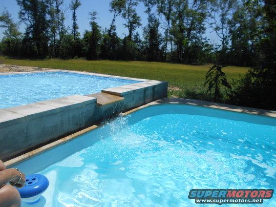 Two-tiered swimming pool with water flowing over the edge, grassy yard and trees in background. Supermotors.net logo in lower right